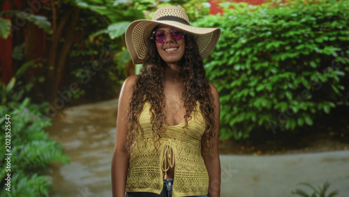Tableau sur toile Hispanic teenage woman smiling in straw sunhat and purple sunglasses, bare shoulders visible, hands relaxed at sides, standing in forest; summer serenity