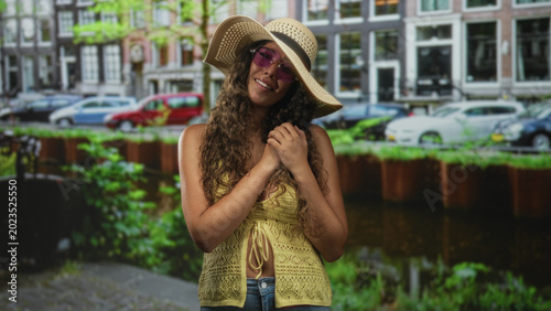 Photos Hispanic woman wearing a straw hat and purple sunglasses, bare midriff visible, hands clasped near chest on a canal street lined with parked cars; carefree summer serenity