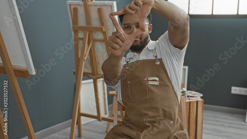 Fotografie Man painter in apron frames scene with hands making a viewfinder in studio; contemplative creativity