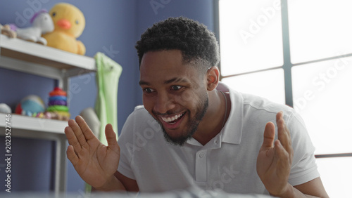 Fotografie Man smiling and clapping hands beside plush duck and toy shelf in a building room, briefly covering his face in playful peekaboo; gentle joy