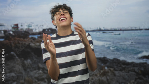 Foto Teenage boy laughing with hands on hips by seaside building, wearing striped shirt and glasses, facing camera; joy playfulness youth