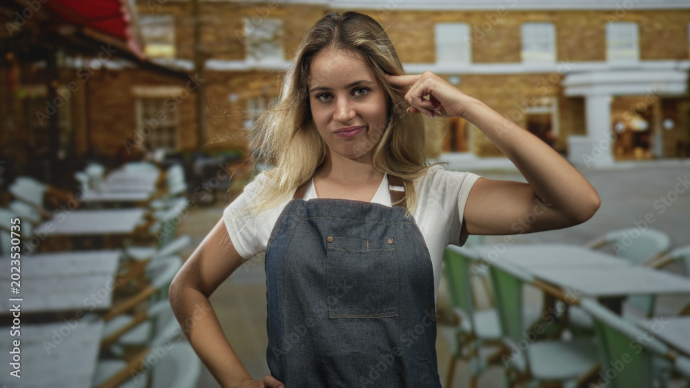 Fototapeta premium Woman pointing finger to temple with one hand on hip on a restaurant terrace by a brick building, wearing denim apron and white t shirt; skepticism.