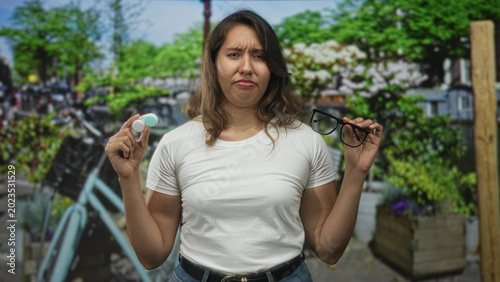 Foto Woman holds a contact lens case in one hand and eyeglasses in the other while standing on a street with a bicycle and planter boxes; annoyance indecision choice dilemma