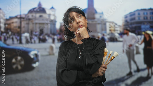 Foto Woman holding makeup brushes with hand on chin in street, city square visible behind her; contemplation creativity