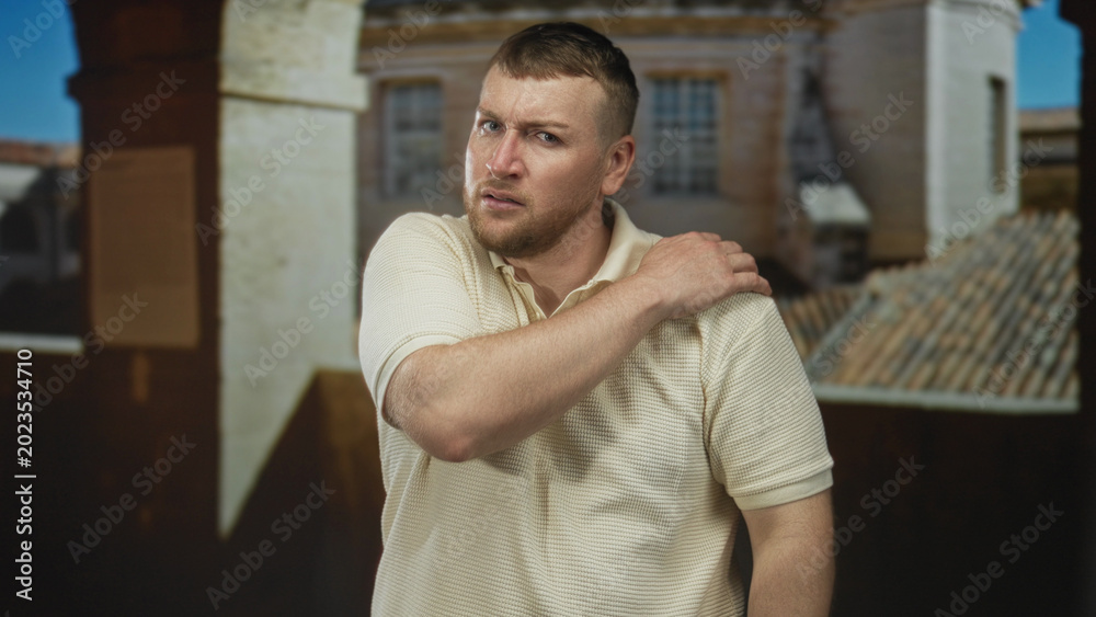 Obraz premium Man clutching left shoulder and wincing, wearing cream polo shirt, standing before an old town building with tiled roof; discomfort concern recovery.