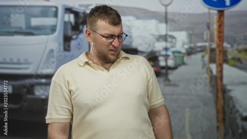Billede på lærred Man grimacing beside parked truck on street wearing glasses and cream polo shirt; disgust unease discomfort