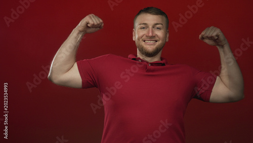 Bilde på lerret Young heavyset man flexing biceps with clenched fists in a red studio set, smiling at the lens; confidence strength