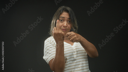 Photography Hispanic woman shows middle finger with left hand while clenching right fist in studio; defiance provocation