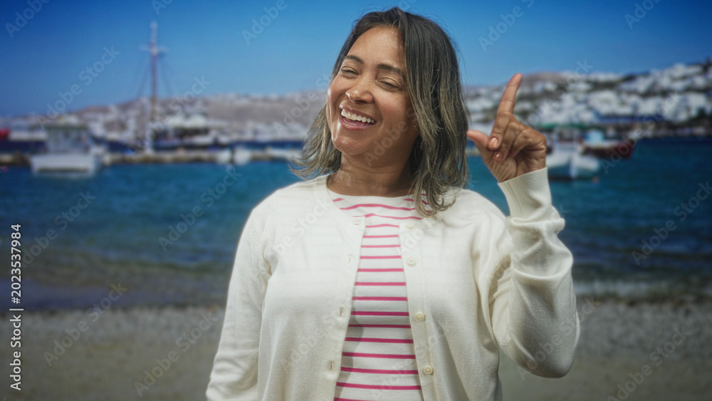 Fototapeta premium Hispanic middleaged woman pointing index finger up, smiling and wearing white cardigan and striped shirt at a harbor; joy playfulness.