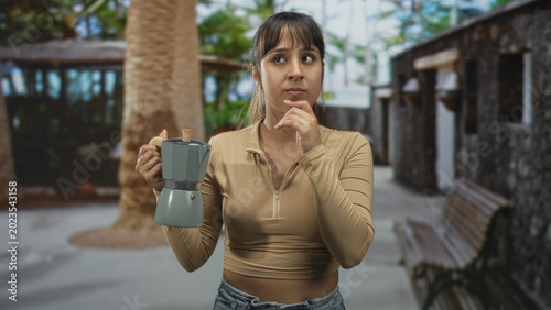 Bild auf Leinwand Young woman holding a moka pot with hand on chin gesture in a stone building courtyard near benches and palm tree; quiet contemplation