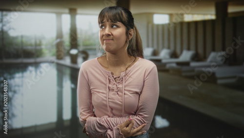 Cuadro en lienzo Young woman with arms crossed at waist beside indoor pool in a building lounge, ponytail and pink longsleeve top, looking sideways; unease hesitation