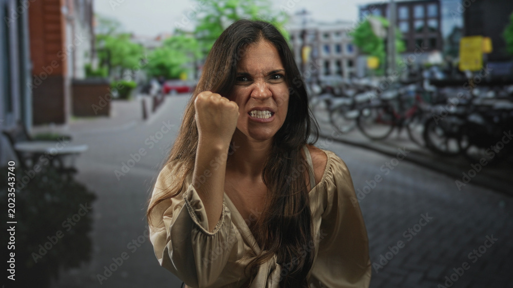 Fototapeta premium Woman with clenched fist raised at camera on city street with bicycles and brick buildings visible; anger defiance resilience.