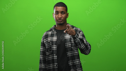 Photos Young black man in plaid shirt points finger to camera in green studio, facing forward with slight head tilt; confidence direct address