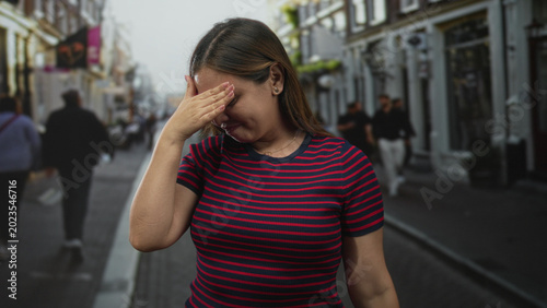 Cuadro en lienzo Woman covers face with both hands on busy city street wearing red striped top and gold necklace while turning head away; shyness privacy