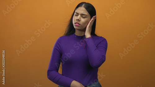 Bild auf Leinwand Woman pressing ear with hand against orange wall in studio setting with pained expression; discomfort