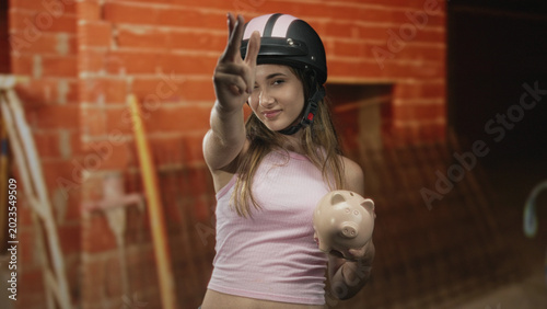 Tableau sur toile Woman holding piggybank points finger like gun toward camera in building with brick wall and ladder; confidence savings youth