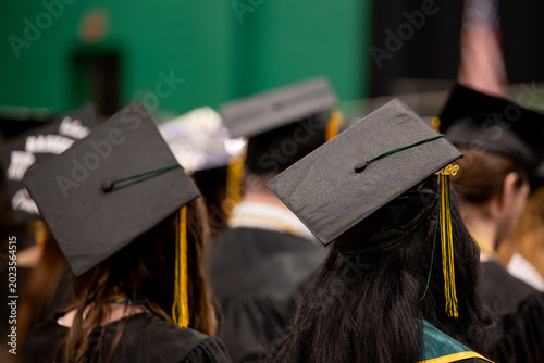 Schilderij op canvas A group of graduates in black gowns and mortarboards with gold tassels are seen from behind at an indoor commencement ceremony, a green gymnasium backdrop behind them