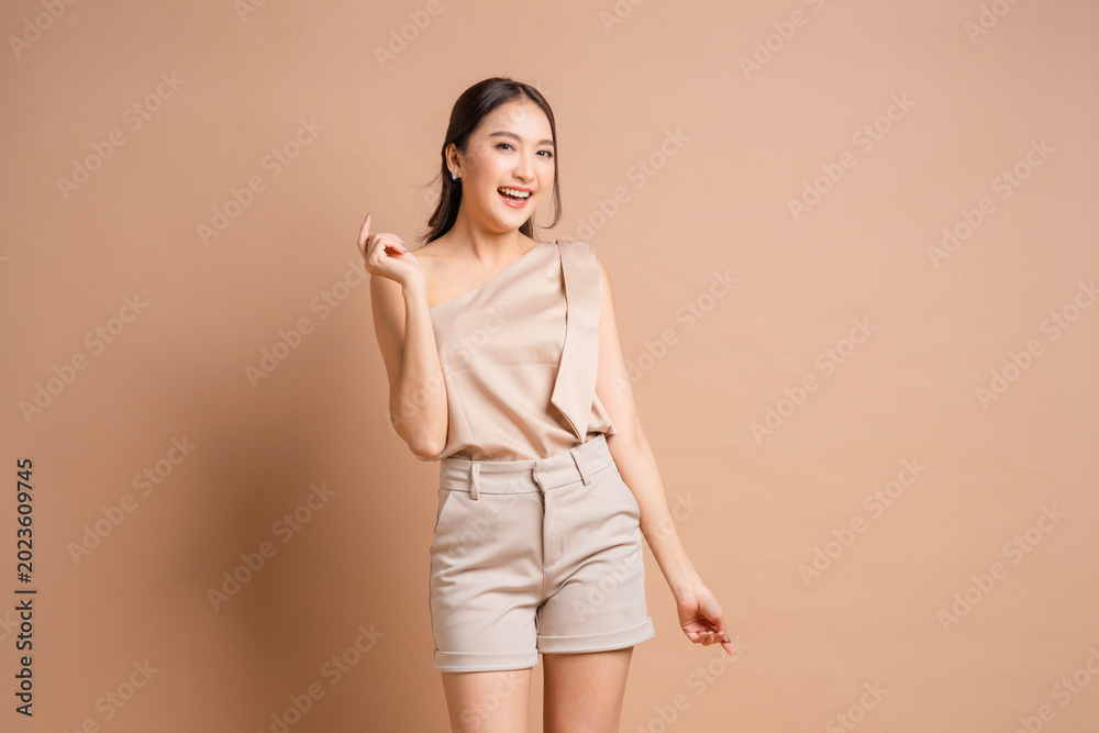 Obraz premium Happy Beautiful Young Asian Woman Pointing Fingers and Looking Away with Shy Excited Smile Portrait of Cheerful Girl in Beige Outfit Posing on Beige Background