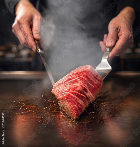 シェフが熱い鉄板で霜降り牛ステーキを焼く様子、ナイフとフォークを使った高級レストランの料理シーン