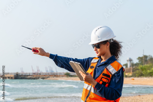 Global offshore maritime industry ESG compliance: Female supervisor using tablet and walkie-talkie to manage coastal logistics operations and corporate CSR environmental sustainability.