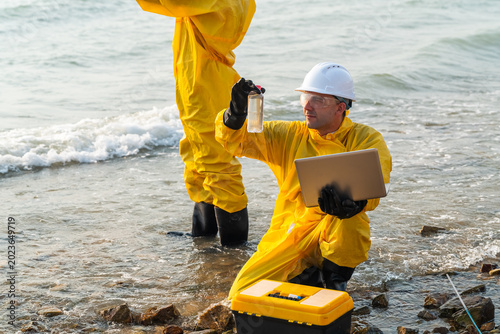Corporate ESG and CSR compliance: Hazmat scientists using laptop for testing toxic ocean water pollution contamination near coastal industrial site for environmental sustainability research.
