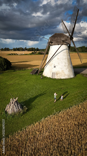 Aerial view of historic Seidla windmill in Estonia with green grass golden wheat fields and a family walking under a dramatic stormy sky shot by DJI Mini 4 Pro drone