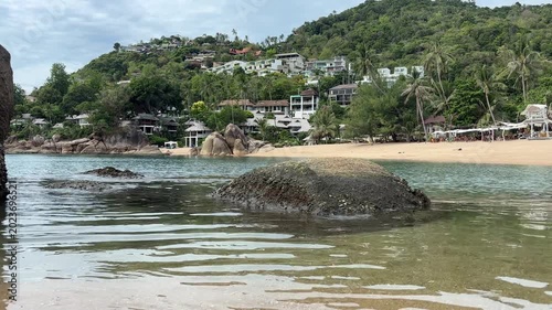 Large rock in clear turquoise water near a tropical beach with white coastal houses and palm trees, showing a peaceful seaside landscape and island atmosphere in Thailand.