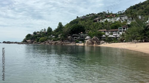 A serene view of a sandy beach and turquoise waters on Samui Island, Thailand. The image showcases a beautiful coastline with rocky formations, beachfront bungalows, and lush green vegetation.