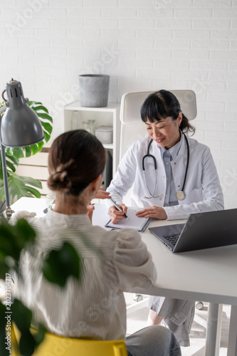 Asian physician writing medical notes during appointment with female patient in bright healthcare office. Preventive care and patient support concept.