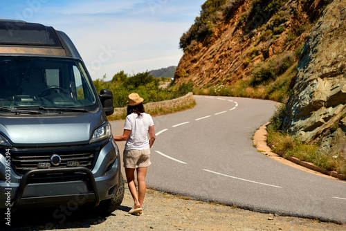 Middle aged woman traveler standing beside a camper van on a winding coastal mountain road during a road trip in Corsica. Colors: gray, blue, beige.