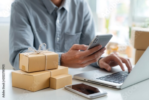 Close up of a small business owner managing online orders. Male entrepreneur using a laptop and smartphone with packages on the desk for shipping