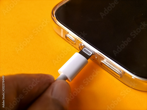 close-up of a person's hands plugging a white charging cable into a smartphone over a vibrant yellow-orange backdrop.