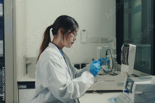 Female scientist in a lab coat and safety glasses carefully weighing white chemical powder in a beaker using a lab scale