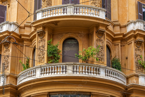 Ornate yellow historical building facade in Rome featuring decorative balcony with plants, intricate architectural details, and closed windows in an urban setting