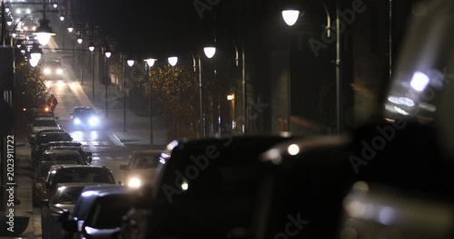  A police car with flashing blue and red lights slowly cruises down a quiet, cobblestone city street at night, while a pedestrian walks by on the side sidewalk.