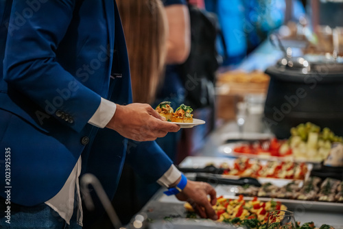 Man in blue blazer holding plate with appetizers at a buffet table featuring various dishes and a blurred background of people and food items