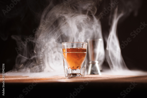 Shot class of dark liquor (whiskey or brandy) centered with dramatic dry ice - smoke creating a custom cocktail appearance. 