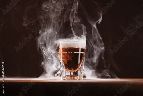 Photography Shot class of dark liquor (whiskey or brandy) centered with dramatic dry ice - smoke creating a custom cocktail appearance