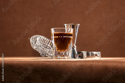 Shot glass of whiskey sitting in front of common stainless steel bartending tools like a strainer, a jigger, and ice tongs. 