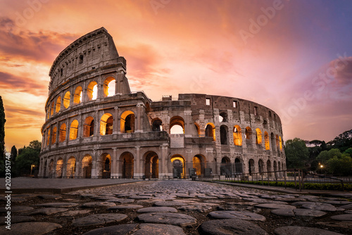 Striking low-angle perspective focusing on the large basalt stone pavers, leading the eye toward the illuminated Colosseum under a golden sunset sky. A premium commercial image combining ancient Roman