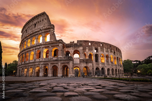 The Colosseum illuminated under a fiery sunset sky with ancient basalt pavement.
