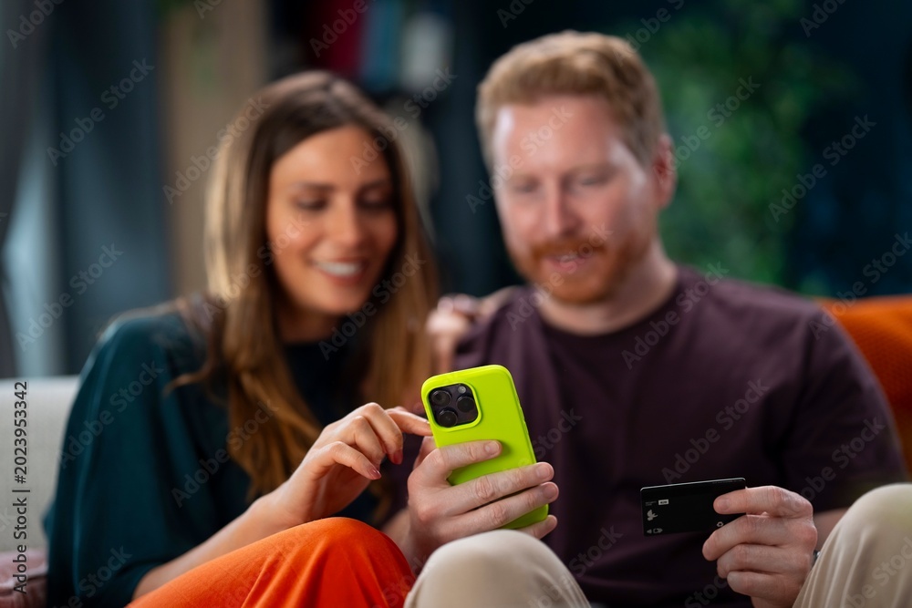Fototapeta premium A man and a woman sit close on a couch. They look at a smartphone and share a credit card. They seem to talk and smile while shopping online in their living room.