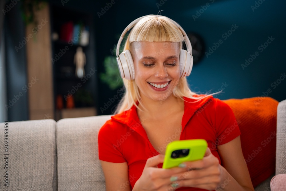 Naklejka premium A woman sits on a couch wearing headphones while looking at her smartphone. She smiles and interacts with her device in a cozy room with colorful decor and soft lighting.