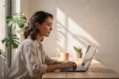 Focused young woman typing on laptop keyboard while working from home in a bright minimalist interior