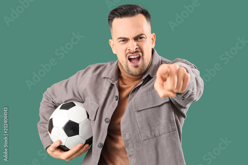 Cuadro en lienzo Angry young man with soccer ball pointing at viewer on green background
