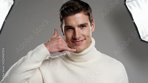 Young Man Gesturing Call Me Sign Wearing White Turtleneck Sweater Against Gray Background Studio Lighting