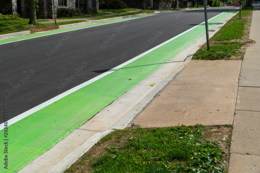 Obraz premium Newly paved asphalt road featuring a vibrant green bike lane with a crisp white stripe, alongside a concrete curb, grassy verge, and sidewalk in a quiet residential area, symbolizing modern urban