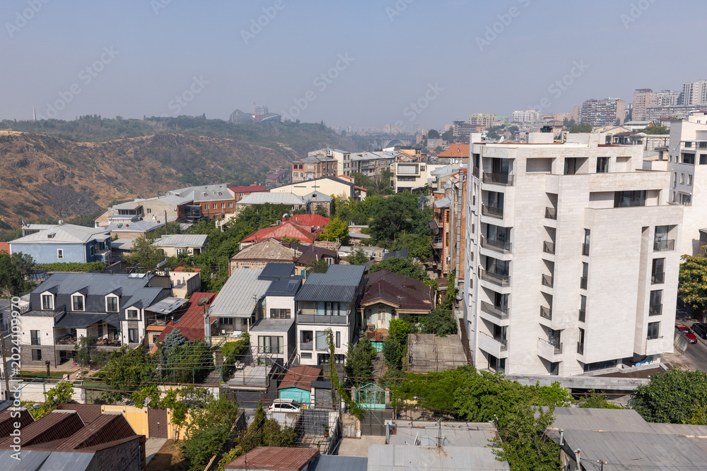 Obraz premium Sunlit urban residential neighborhood with mixed traditional houses and a modern apartment building on a hillside, showing rooftops, narrow streets, greenery and a city skyline of Yerevan, Armenia