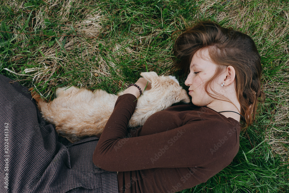 Fototapeta premium Woman lies on grass cuddling a dog, cozy outdoor moment in a brown sweater, natural light, relaxed lifestyle scene, affectionate bond, peaceful mood and quiet countryside feeling.