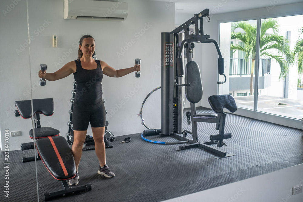 Fototapeta premium Young woman in black outfit performing dumbbell exercises with arms extended in a bright modern gym with large windows and equipment. Active fitness concept
